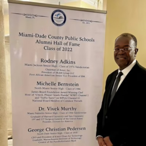 A man standing next to a sign that reads " miami dade county public schools alumni hall of fame class 2 0 1 2 ".